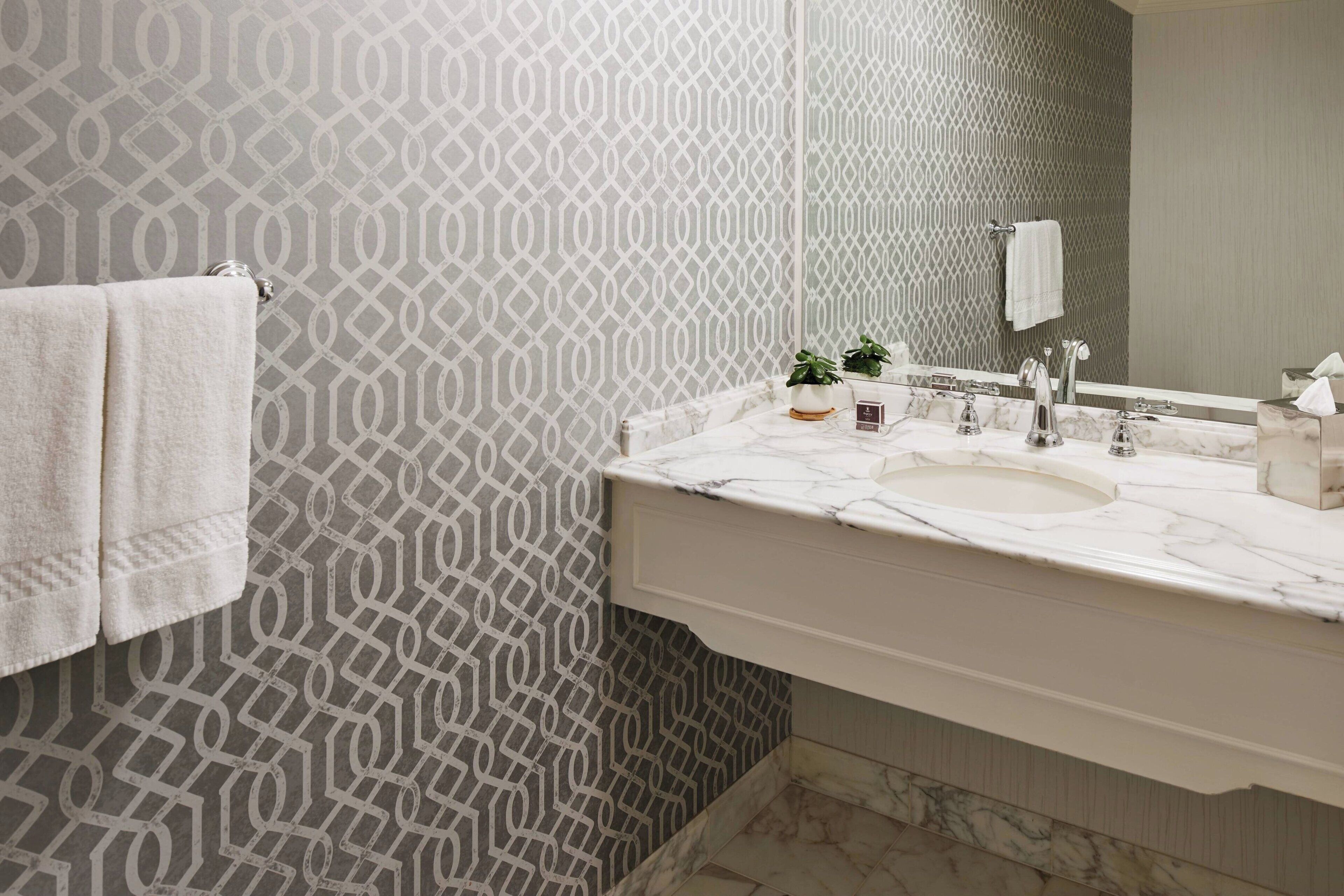 A bathroom with a patterned wallpaper, a white sink, a mirror, and a towel rack.