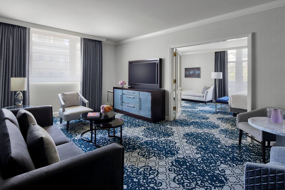 A hotel room with a living area featuring a sofa, a coffee table, a TV, and a cabinet. There is a patterned carpet and a view of another room through an open door.