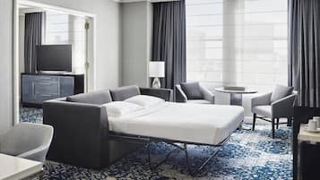 A hotel room with a bed, two armchairs, a small table, a TV, and a minibar.
