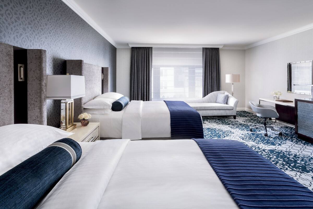 A hotel room with two beds, a desk, a chair, and a large window with curtains.