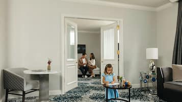 A hotel room with a round table, a sofa, a small table with a vase, and a child playing.