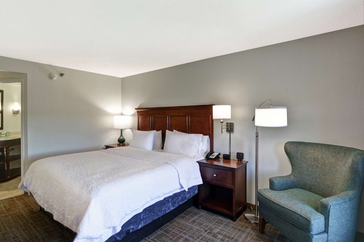 Ein Hotelzimmer mit einem großen Bett, Nachttischlampen, einem Nachttisch, einem grünen Sessel und einem im Hintergrund sichtbaren Badezimmer.