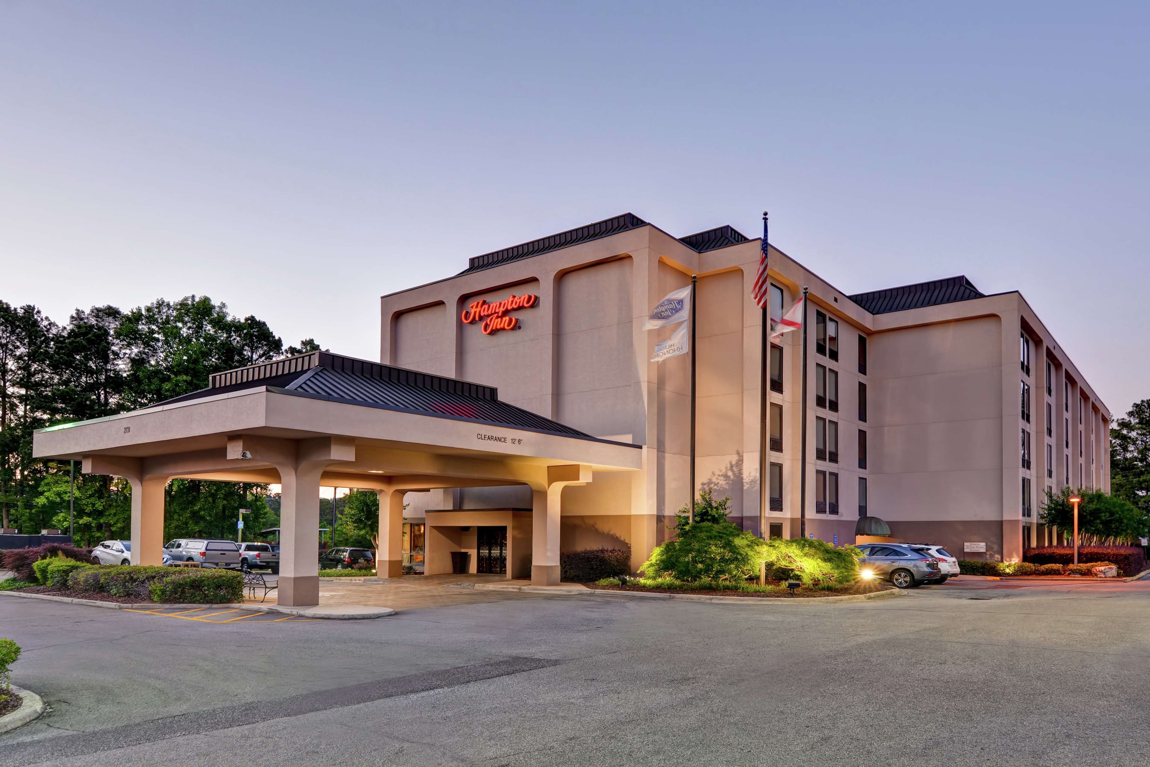 A Hampton Inn hotel with a covered entrance, parking area, and a flagpole with an American flag.