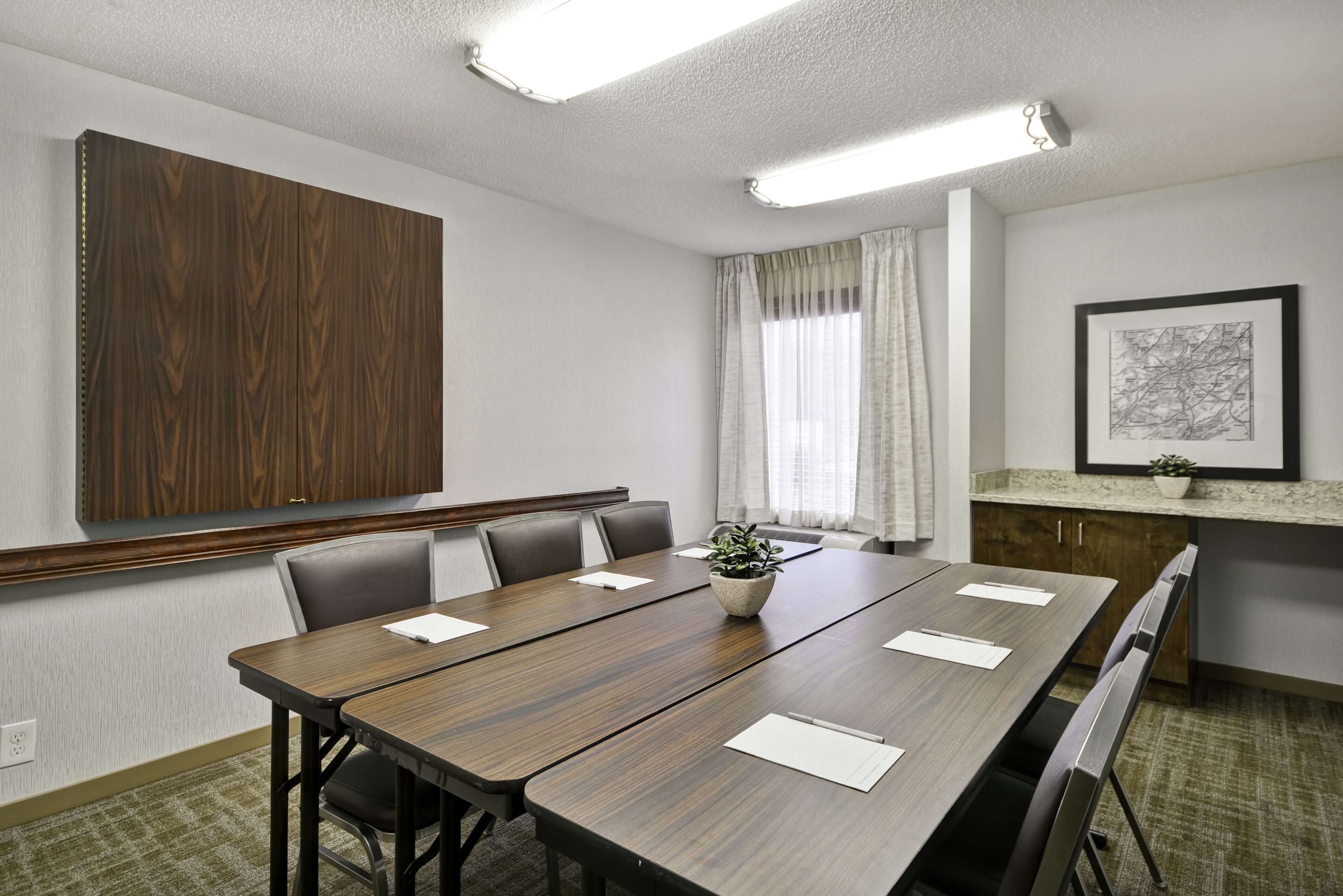 Un salón de conferencias con una larga mesa, sillas, una planta, una obra de arte enmarcada y una ventana con cortinas.