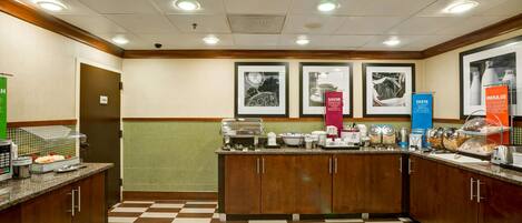 A hotel kitchen with a checkered floor, a buffet counter with various food items, and framed pictures on the wall.