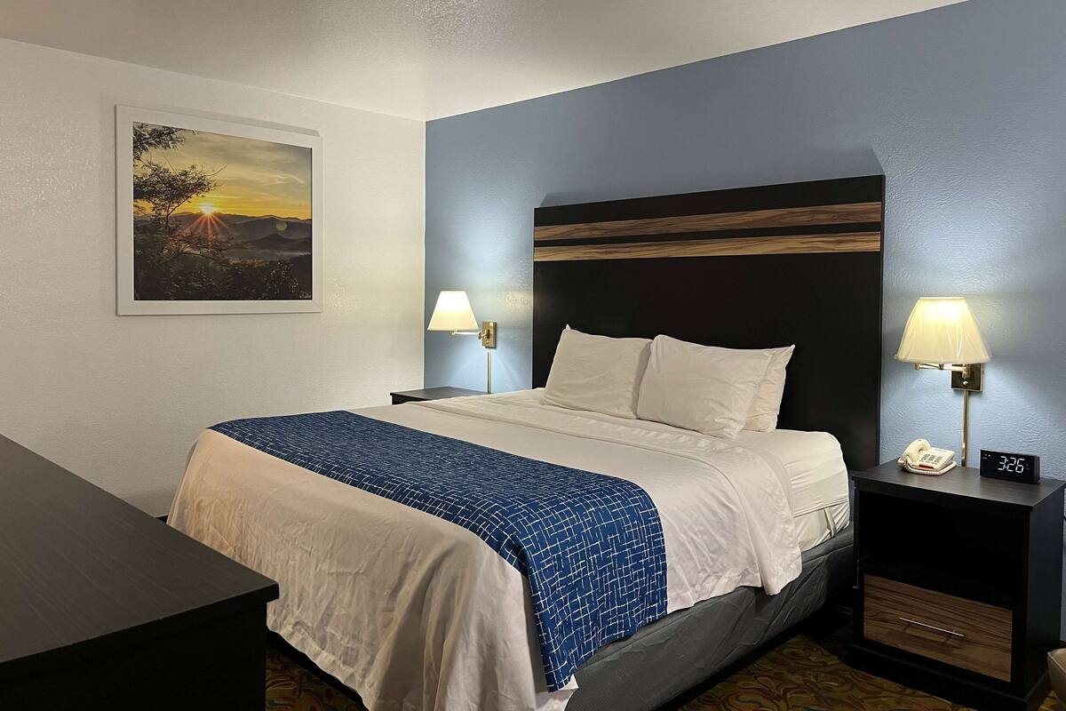 A hotel room with a bed, two bedside tables, a nightstand, a wall-mounted lamp, a painting, and a digital clock.