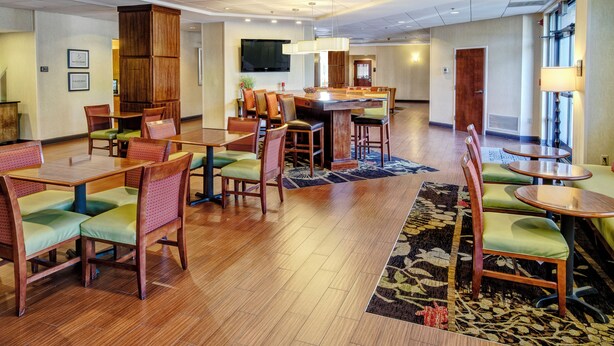 A hotel lobby with wooden flooring, green cushioned chairs, a central bar area, and a television mounted on the wall.