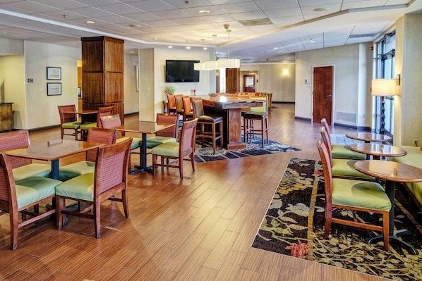 A hotel lobby with wooden flooring, green cushioned chairs, a central bar area, and a television mounted on the wall.