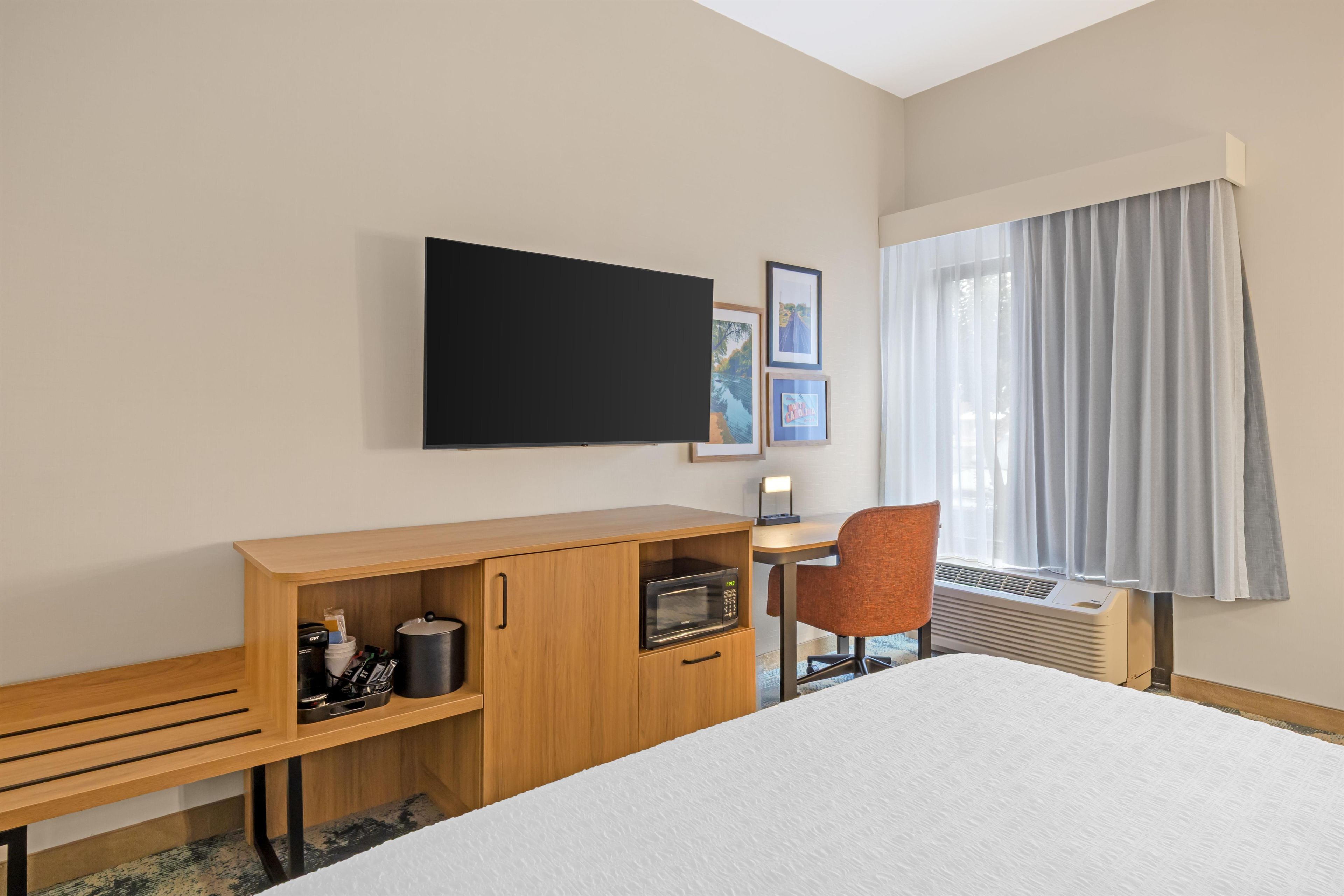 Habitación de hotel con cama, escritorio con silla, televisión de pantalla plana, microondas, calefacción y ventana con cortinas.