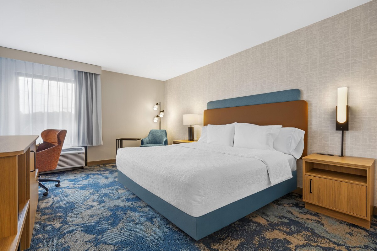 A hotel room with a large bed, a desk, a chair, and a desk lamp.
