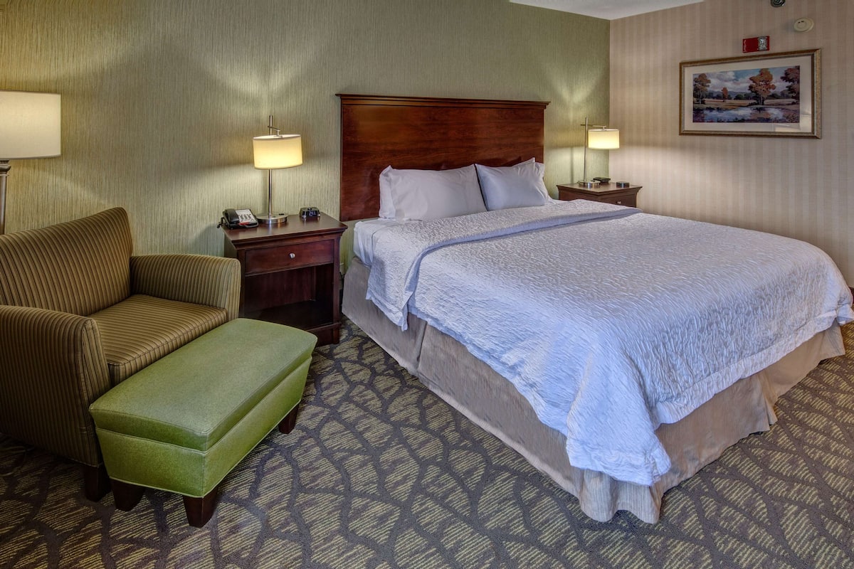 Hotelkamer met een groot bed, een groene bekleden bank, een houten hoofdeinde, twee nachtkastjes met lampen, en een lijst met een afbeelding aan de muur.