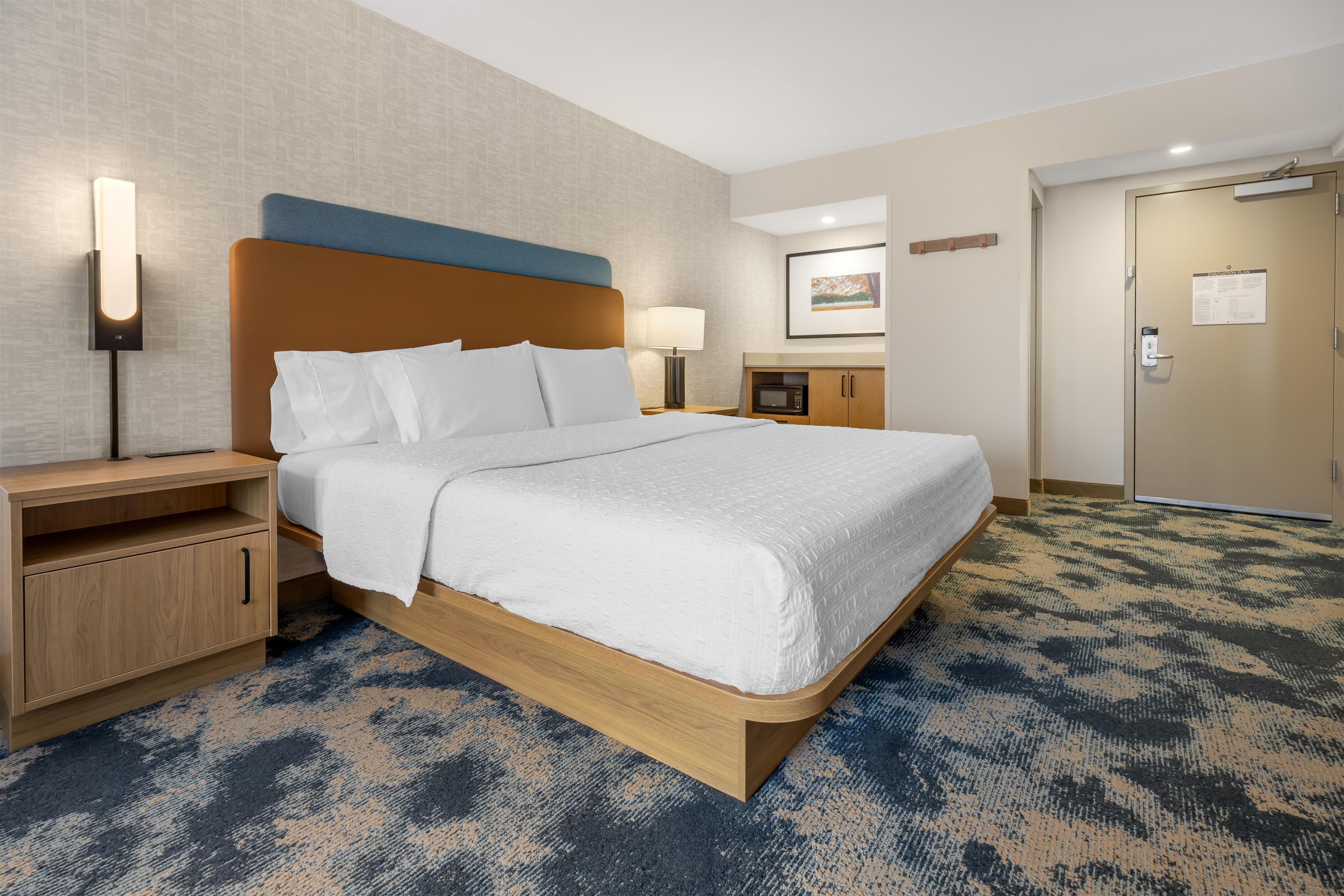 Una habitación de hotel con cama, mesita de noche, lámpara y microondas.