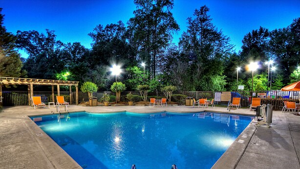 A rectangular swimming pool with a wooden deck and orange chairs, surrounded by trees and illuminated by street lamps.