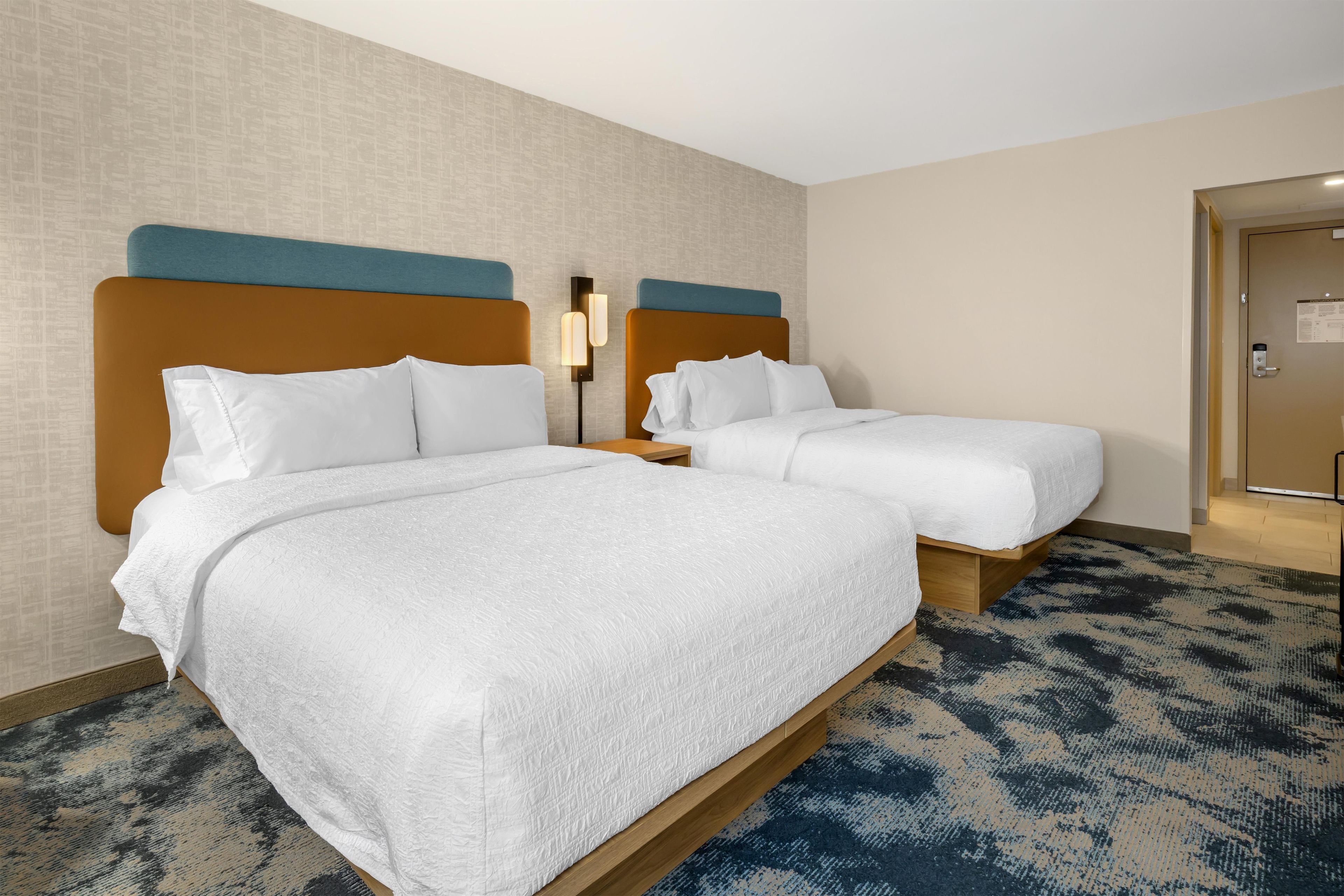 A hotel room with two beds, a nightstand, a wall-mounted lamp, and a door with a lock.