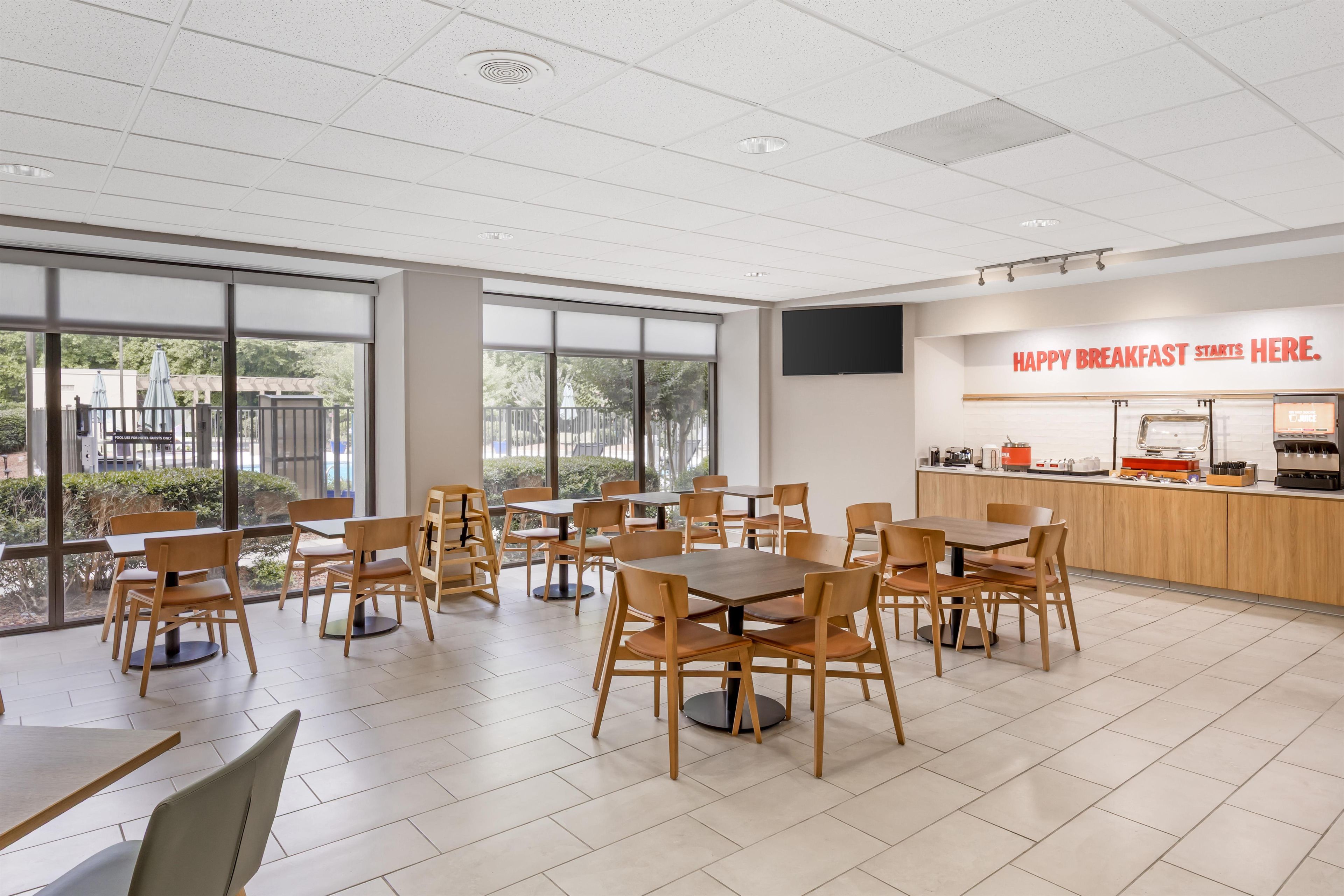 Un comedor moderno con mesas y sillas de madera, ventanas grandes y una barra de cocina con un letrero que dice 'HAPPY BREAKFAST STARTS HERE'.