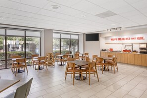 A modern dining area with wooden tables and chairs, large windows, and a kitchen counter with a sign reading 'HAPPY BREAKFAST STARTS HERE.' - Hampton Inn Rocky Mount (Rocky Mount)
