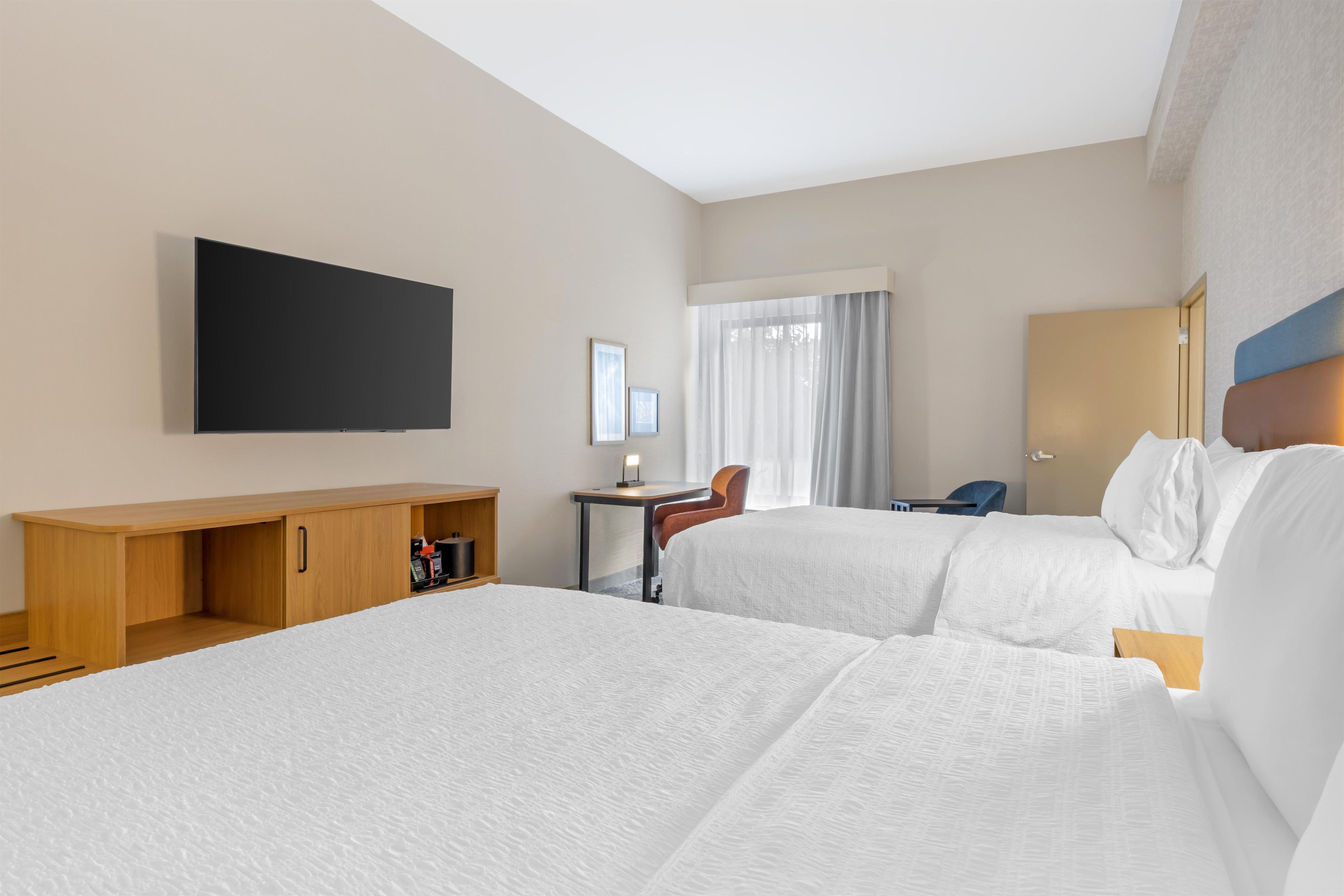 A hotel room with two beds, a TV mounted on the wall, a desk with a chair, and a window with curtains.