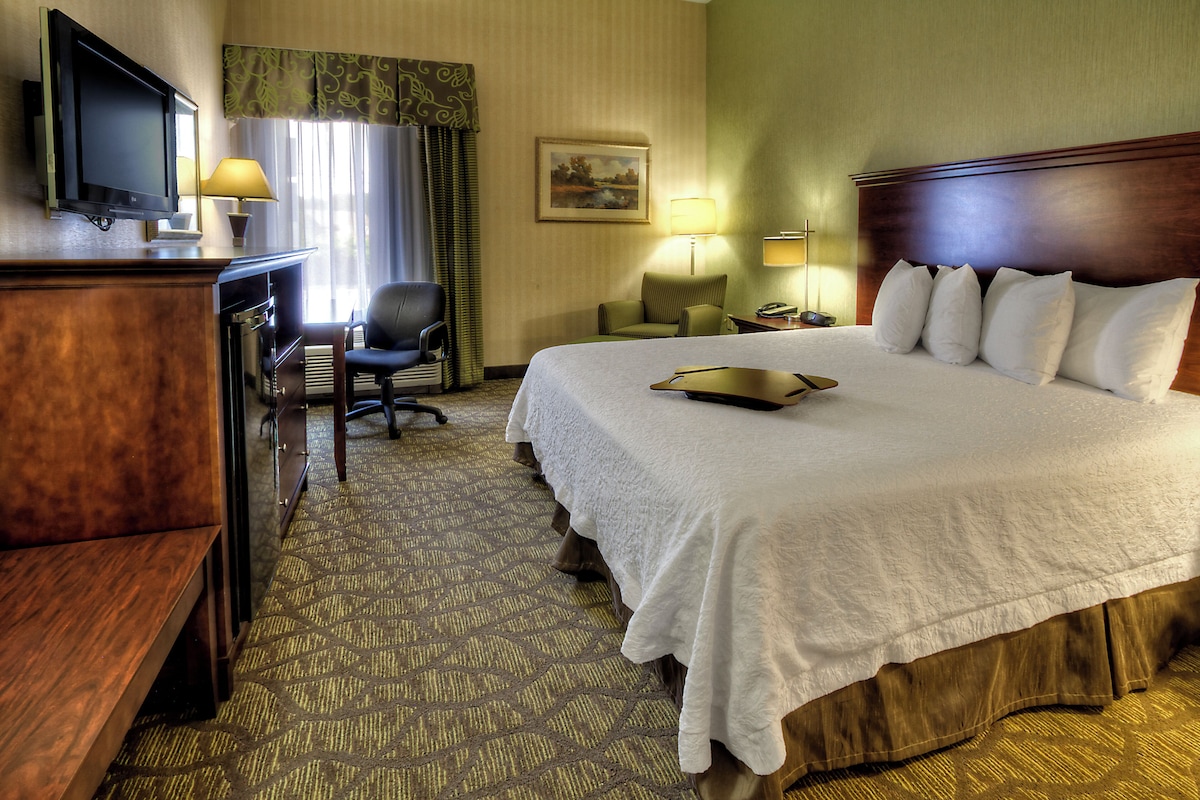 A hotel room with a large bed, a desk with a television, a chair, and a lamp.