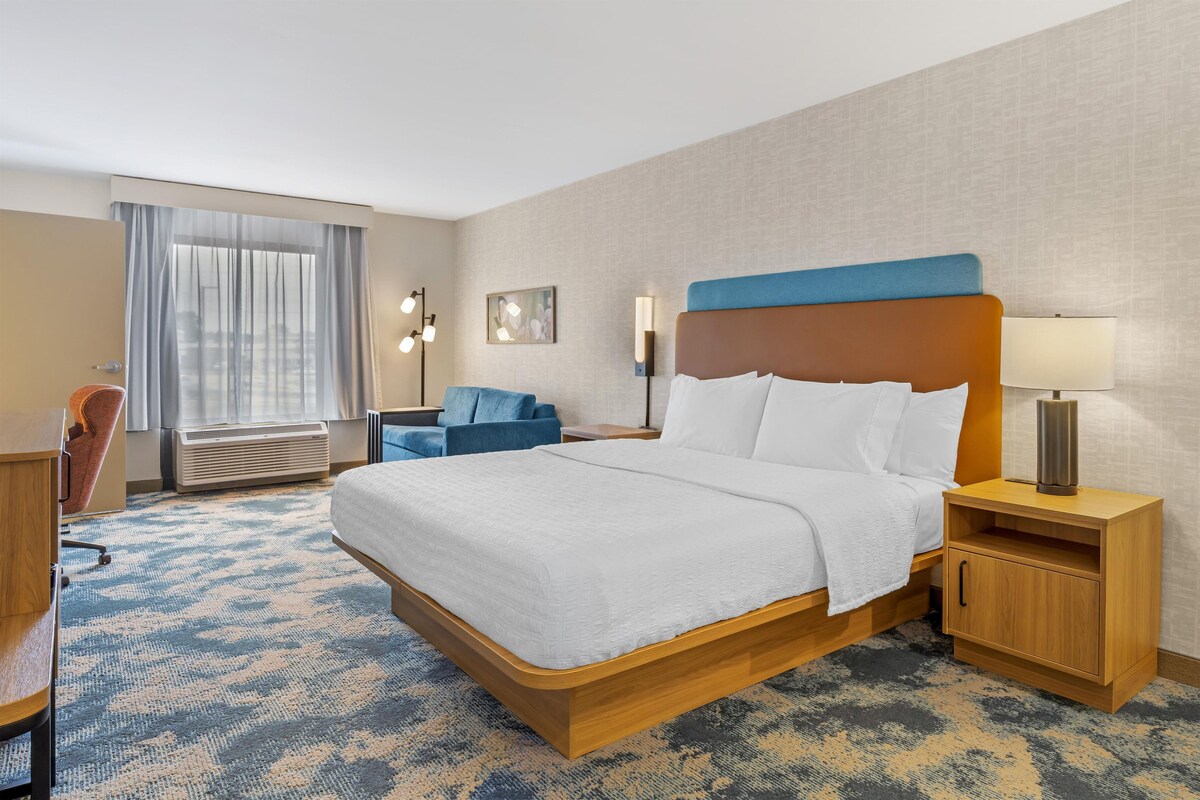 A hotel room with a large bed, a desk, a chair, a bedside table, and a lamp.