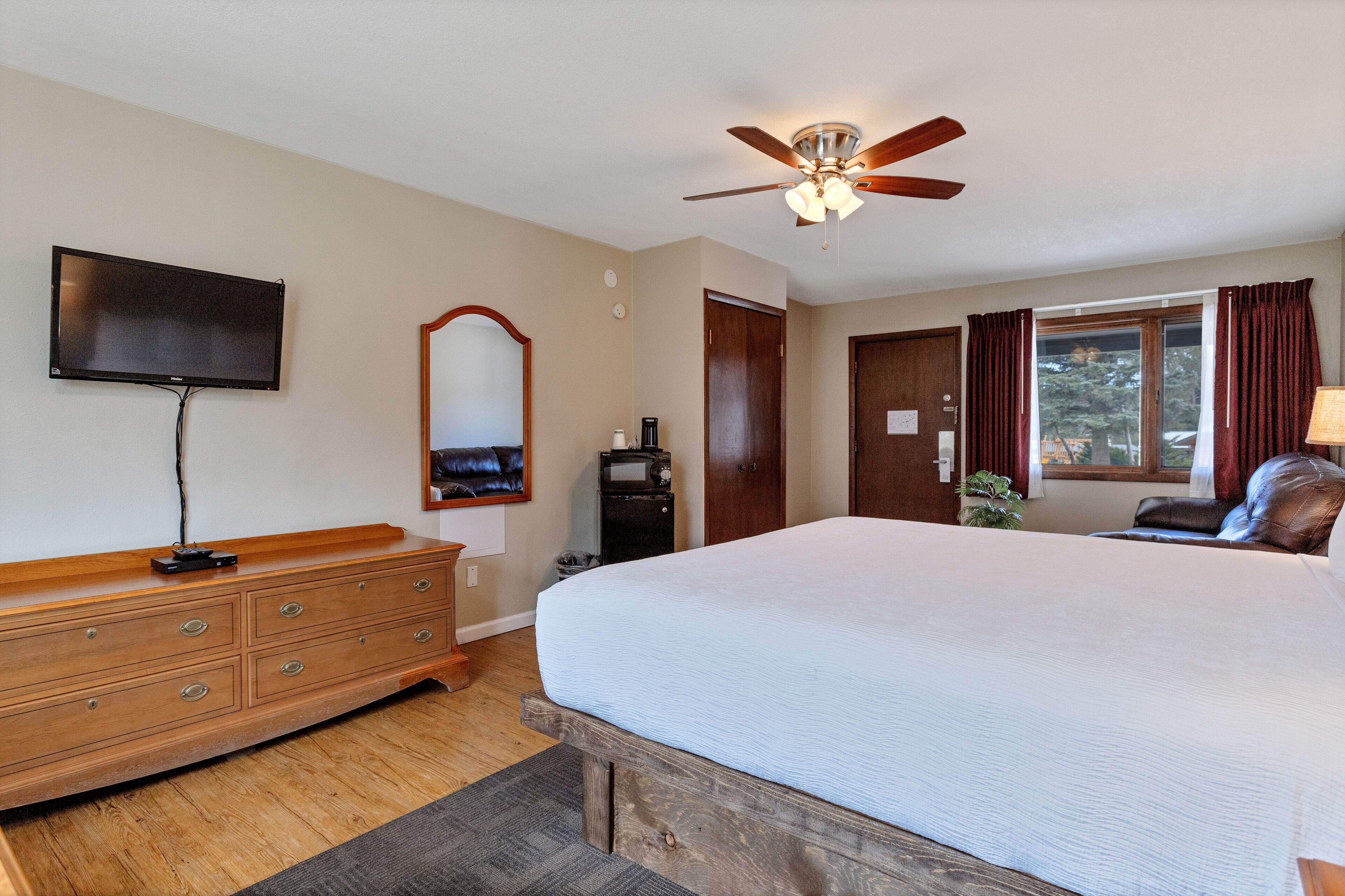 A bedroom with a large bed, wooden dresser, ceiling fan, TV, and a window with curtains.