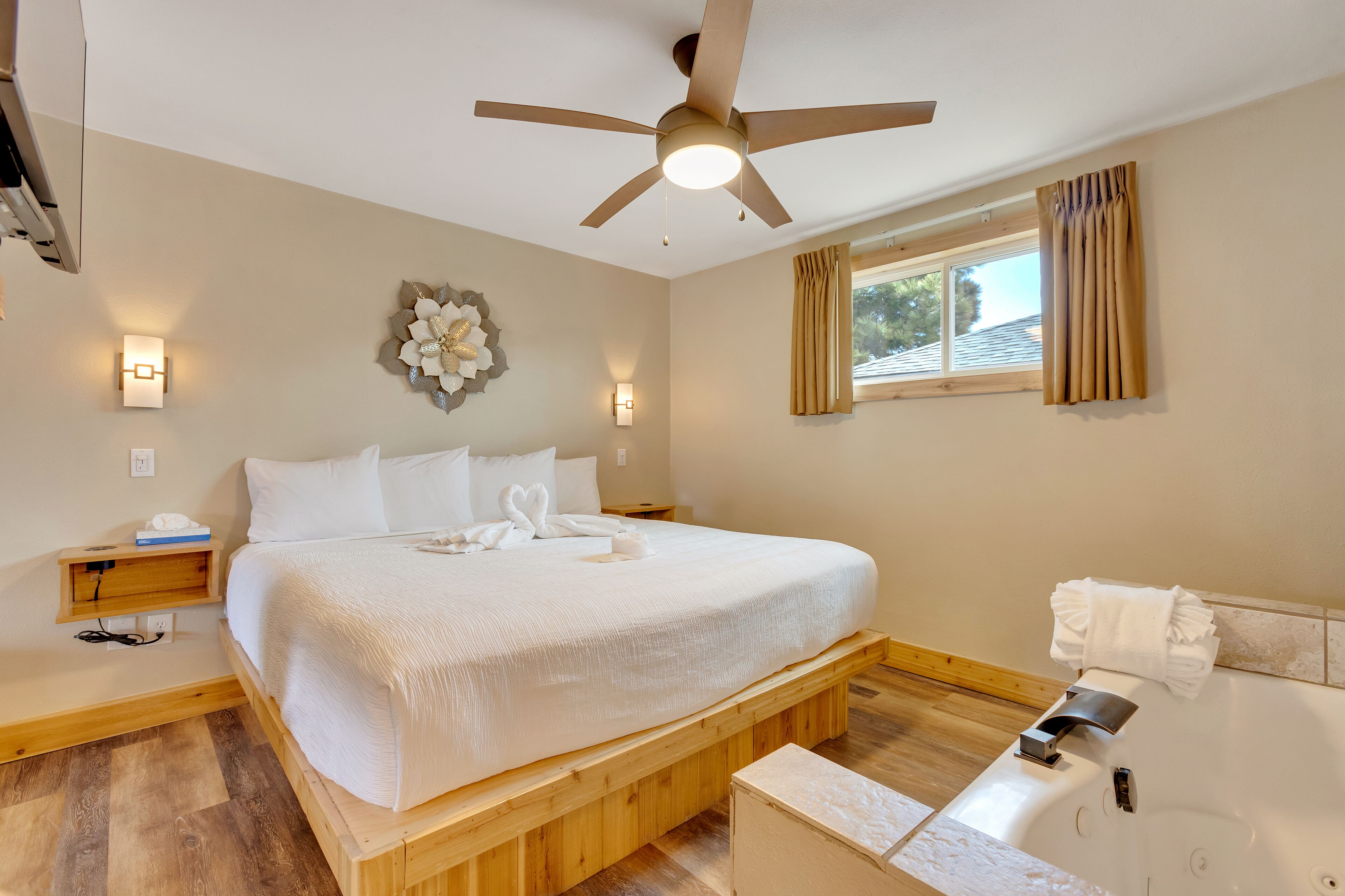 Une chambre avec un grand lit, un ventilateur de plafond, une table de chevet, une commode, une lampe fixée au mur, une fenêtre avec des rideaux et une salle de bain avec une baignoire et un porte-serviettes.