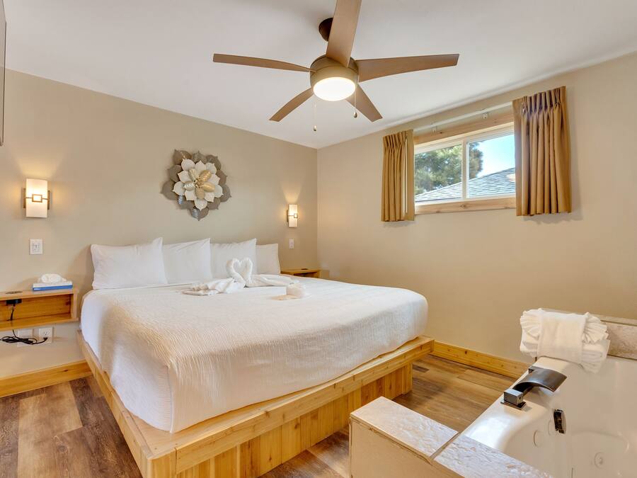 Une chambre avec un grand lit, un ventilateur de plafond, une table de chevet, une commode, une lampe fixée au mur, une fenêtre avec des rideaux et une salle de bain avec une baignoire et un porte-serviettes.