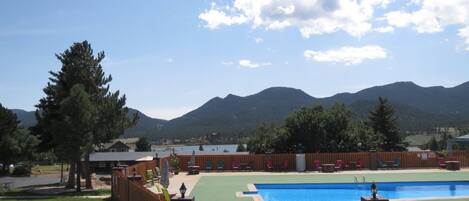 Una piscina con cerramiento de madera, rodeada de césped y árboles, con montañas al fondo.