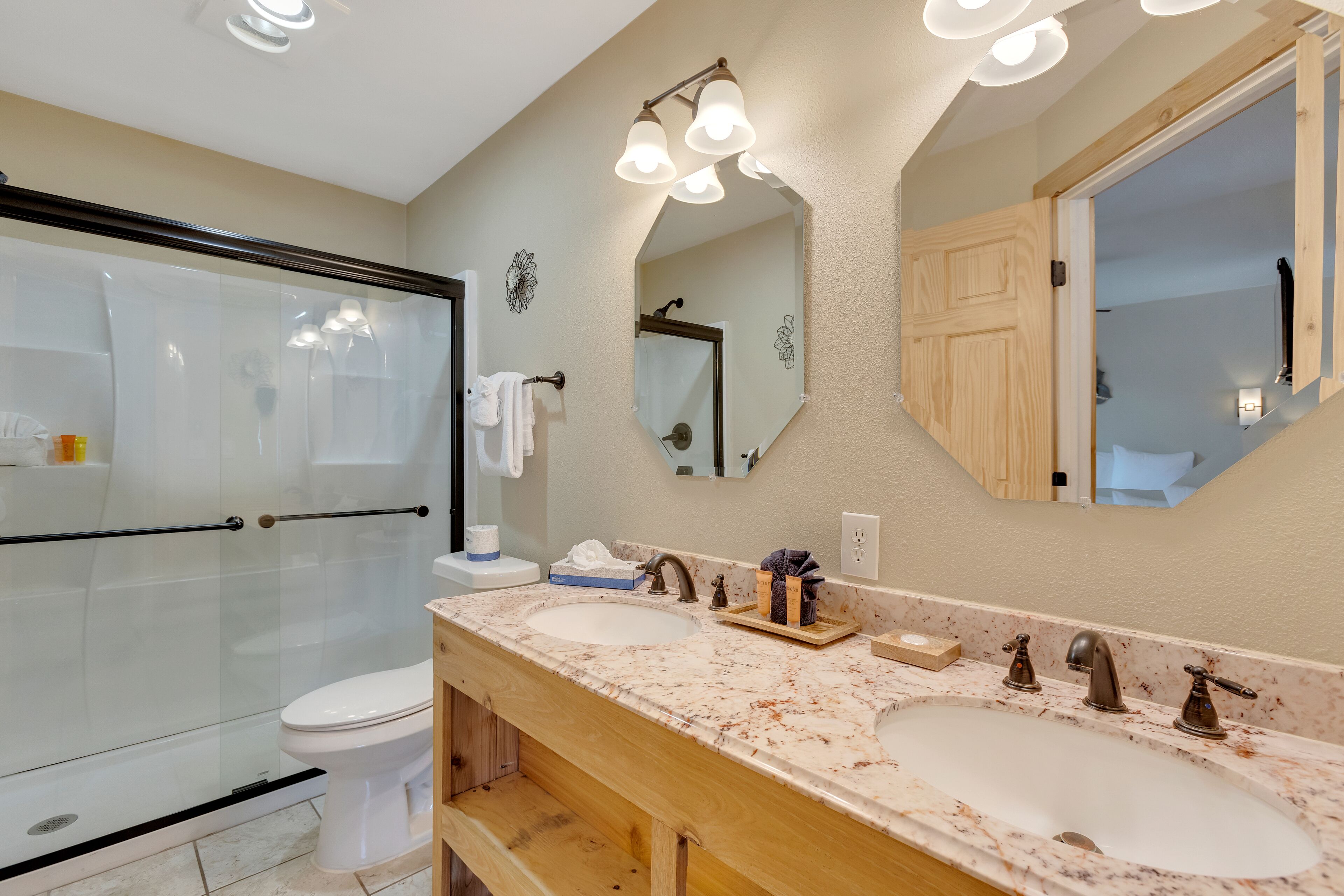 A bathroom with a glass shower enclosure, a toilet, a double sink with a marble countertop, and a large mirror.