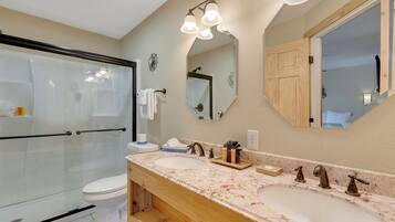 A bathroom with a glass shower enclosure, a toilet, a double sink with a marble countertop, and a large mirror.