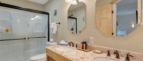 A bathroom with a glass shower enclosure, a toilet, a double sink with a marble countertop, and a large mirror.