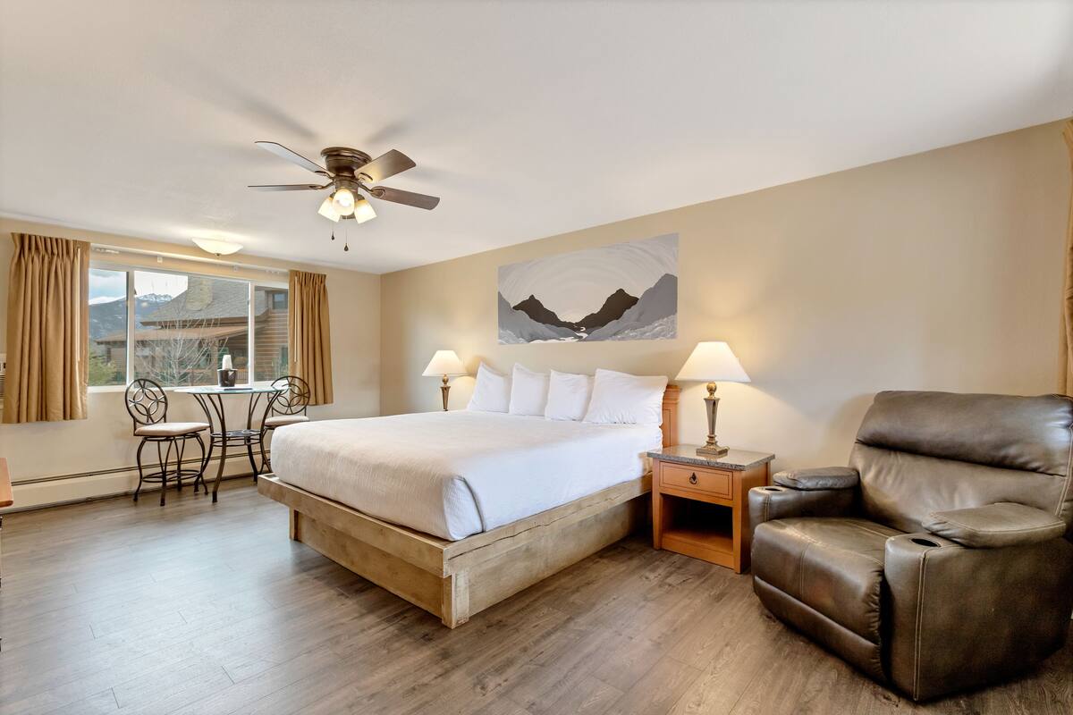 A hotel room with a bed, a reclining chair, a desk with two chairs, a ceiling fan, and a window with curtains.