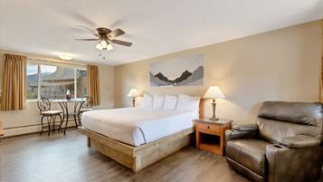 A hotel room with a bed, a reclining chair, a desk with two chairs, a ceiling fan, and a window with curtains.