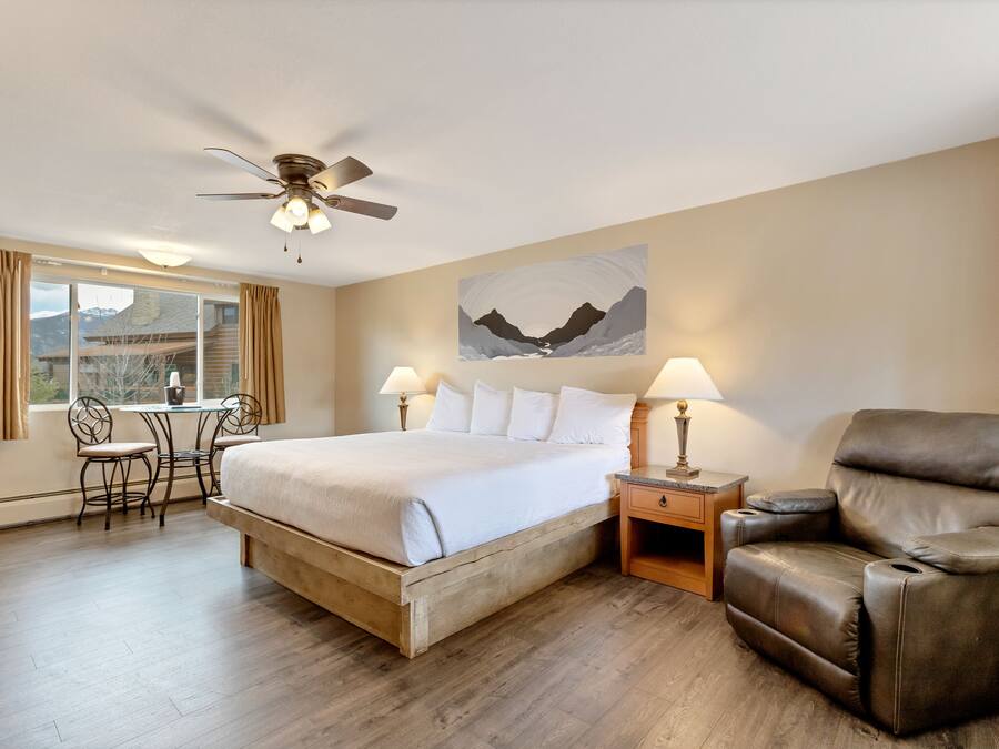 A hotel room with a bed, a reclining chair, a desk with two chairs, a ceiling fan, and a window with curtains.