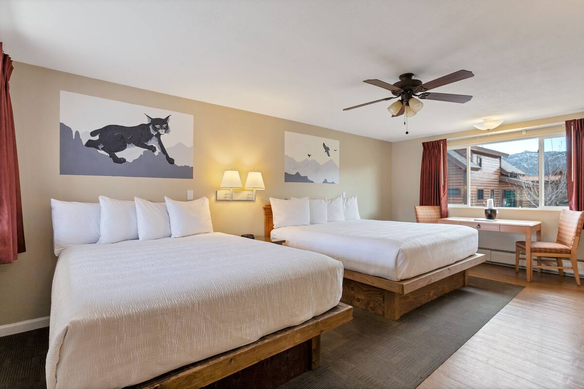 A hotel room with two beds, a desk, a chair, a ceiling fan, and artwork on the walls.
