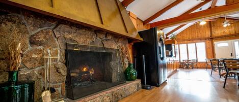 A stone fireplace with a modern design, a wooden ceiling with exposed beams, and a dining area with wooden chairs and tables.