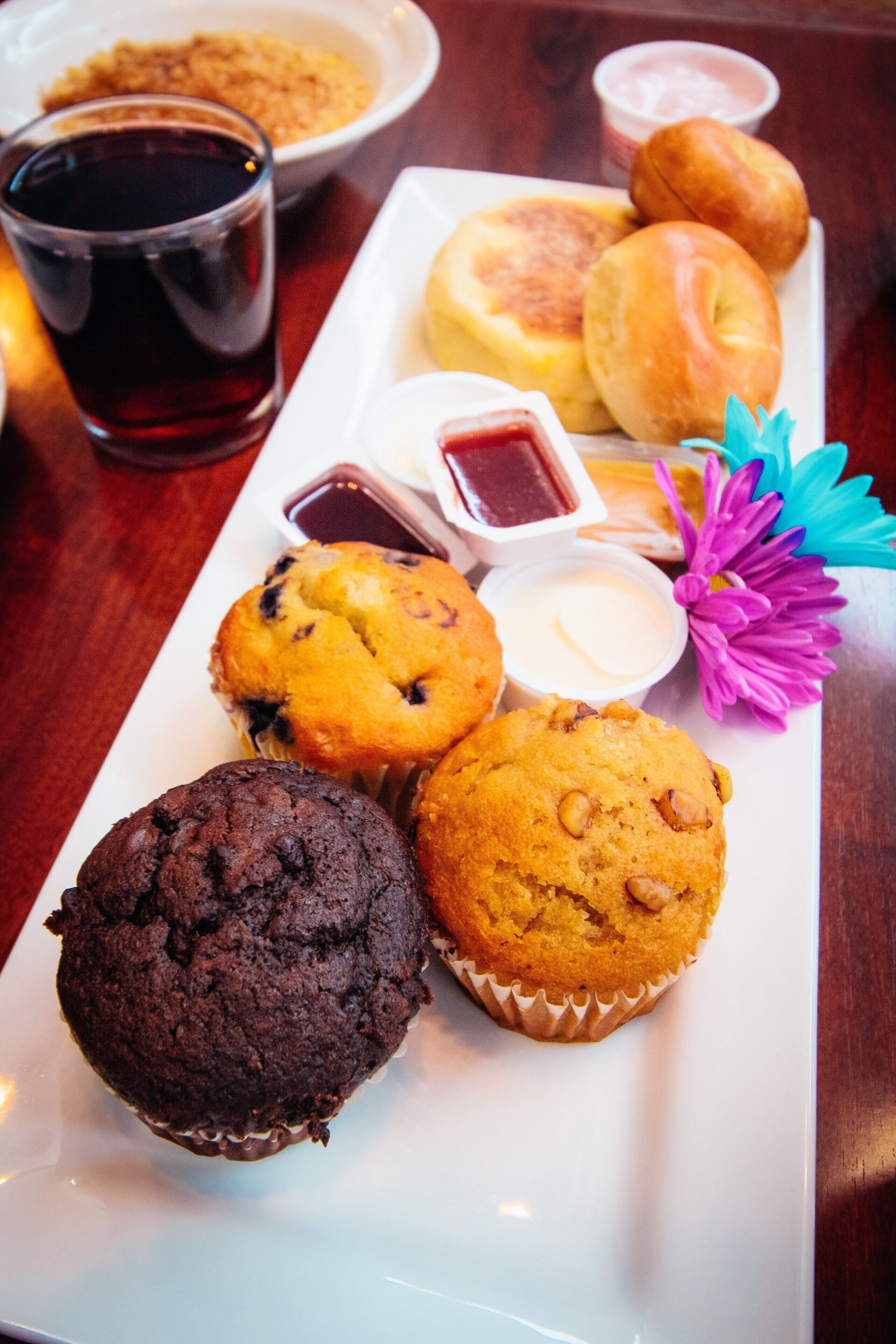 A tray of muffins and pastries on a white plate, accompanied by a glass of dark liquid and small containers of dipping sauces.