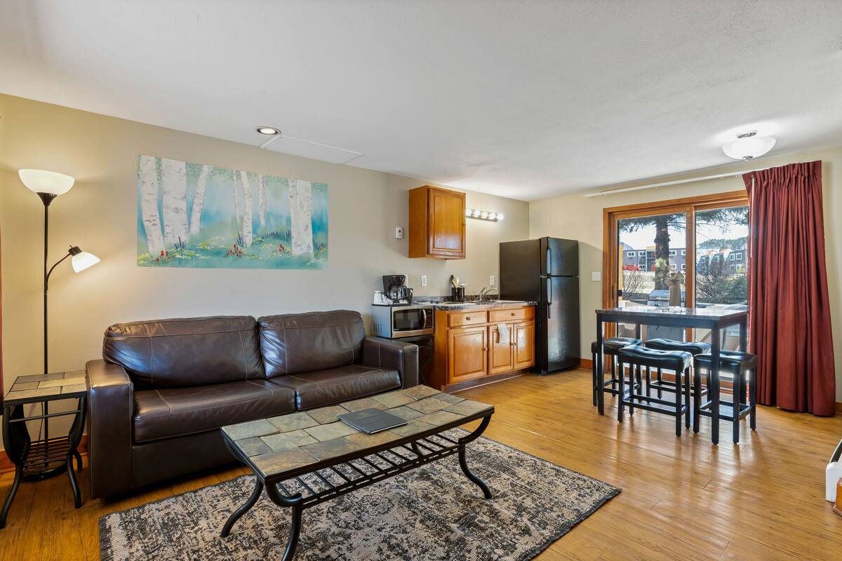 A living room with a brown leather sofa, a coffee table, a painting of birch trees, a kitchen area with wooden cabinets, a refrigerator, and a dining table with chairs.