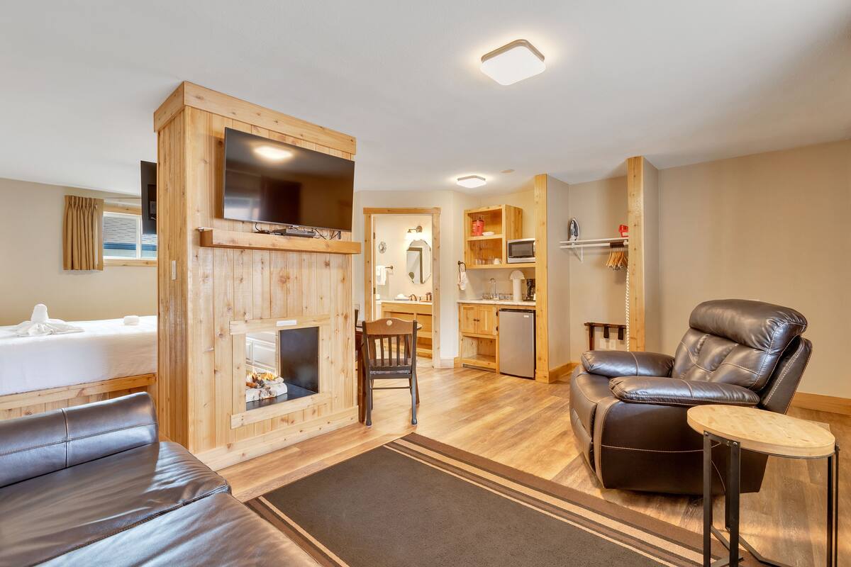 A modern room with a sofa, a recliner, a TV, a fireplace, and a kitchenette.