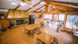 Une salle à manger spacieuse, avec des tables et des chaises en bois, une cheminée en pierre et de grandes fenêtres donnant sur l’extérieur.