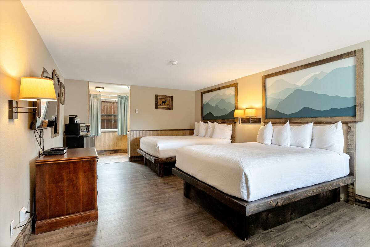 A hotel room with two beds, a wooden dresser, a desk with a lamp, and a painting of mountains.