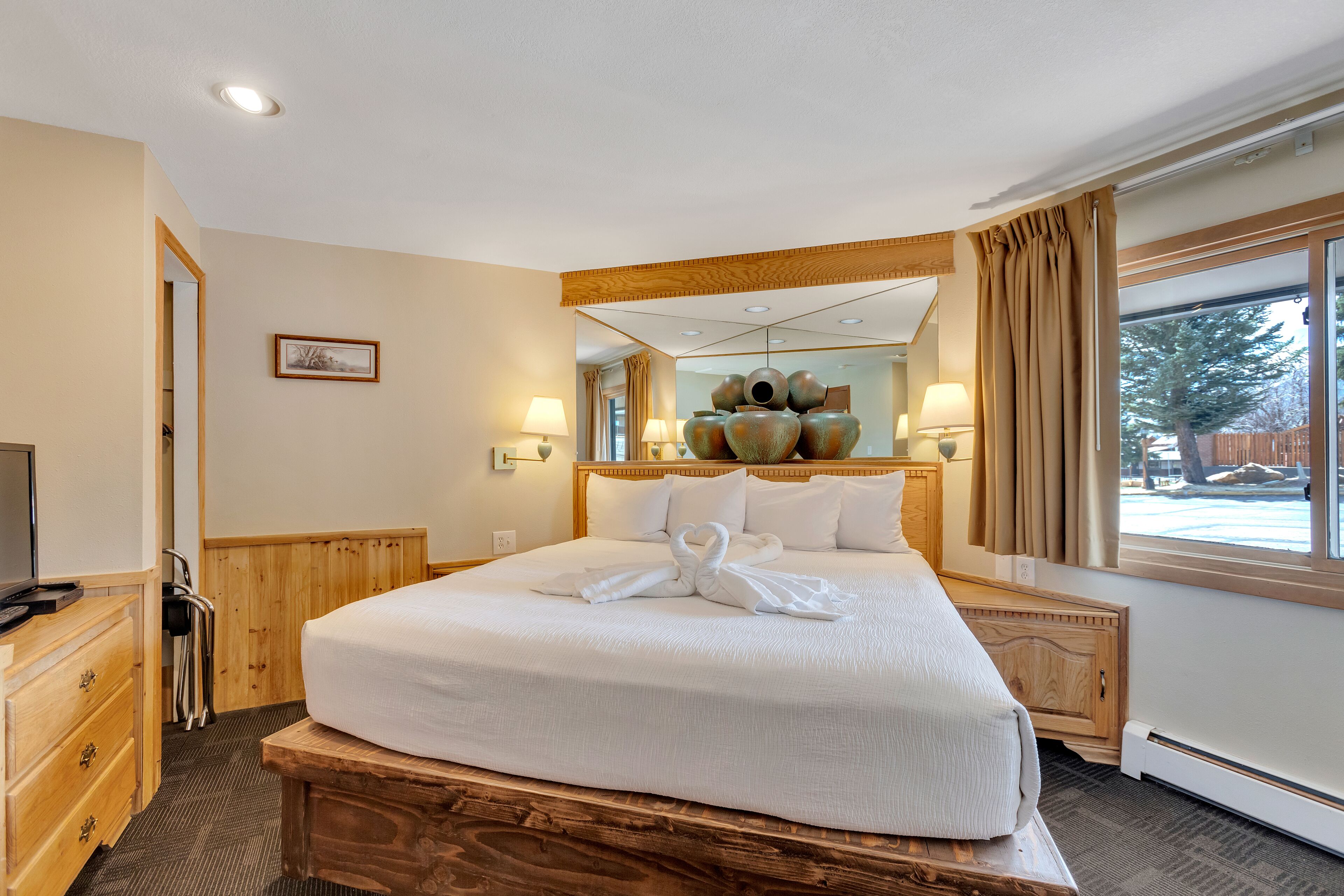 A hotel room with a large bed, wooden headboard, bedside lamps, a television, and a window with curtains.