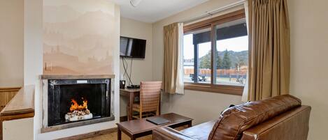 A living room with a fireplace, a sofa, a table, a chair, a TV mounted on the wall, and a window with curtains.
