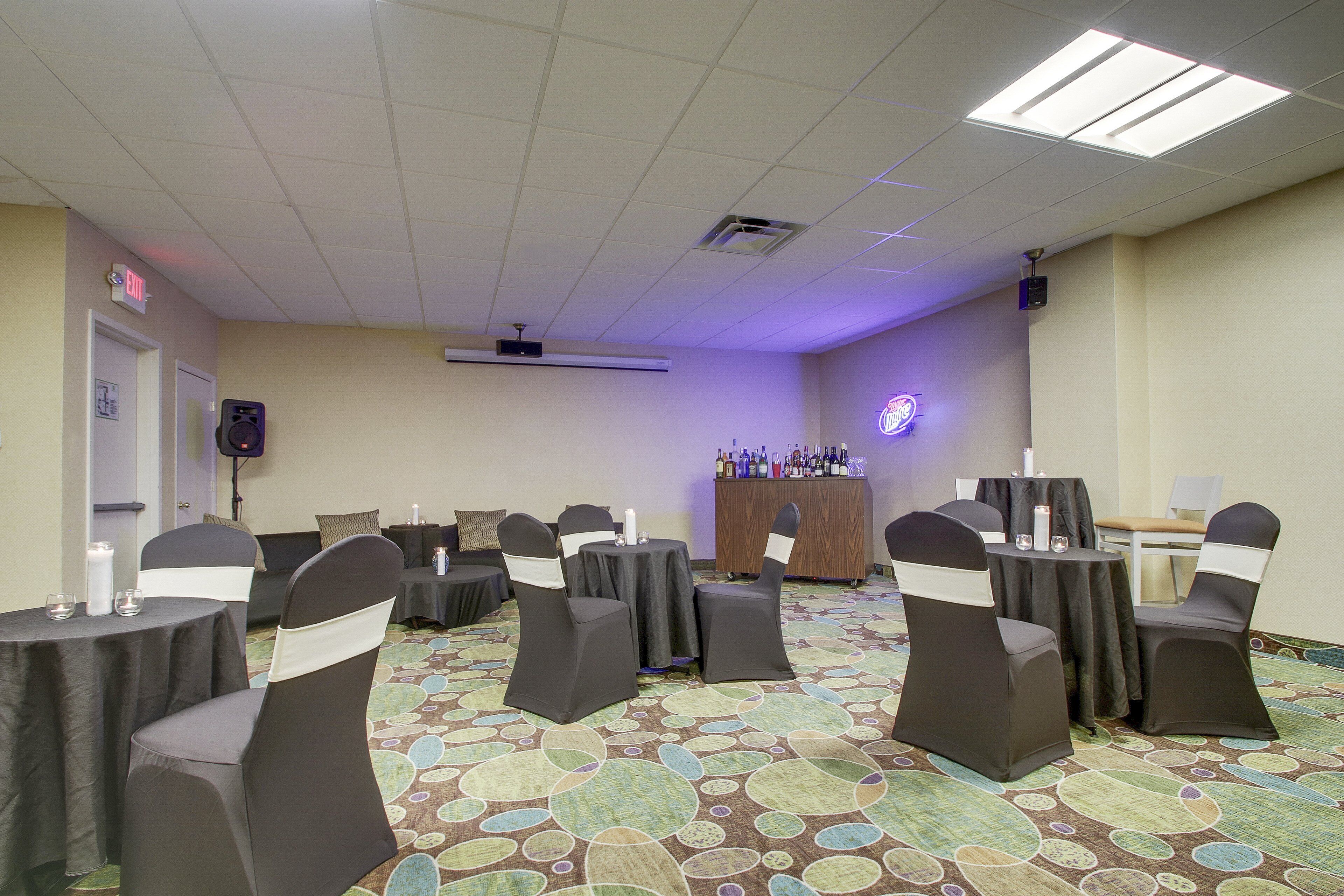 A conference room with round tables, black chairs with white sashes, a podium, and a bar area with bottles.