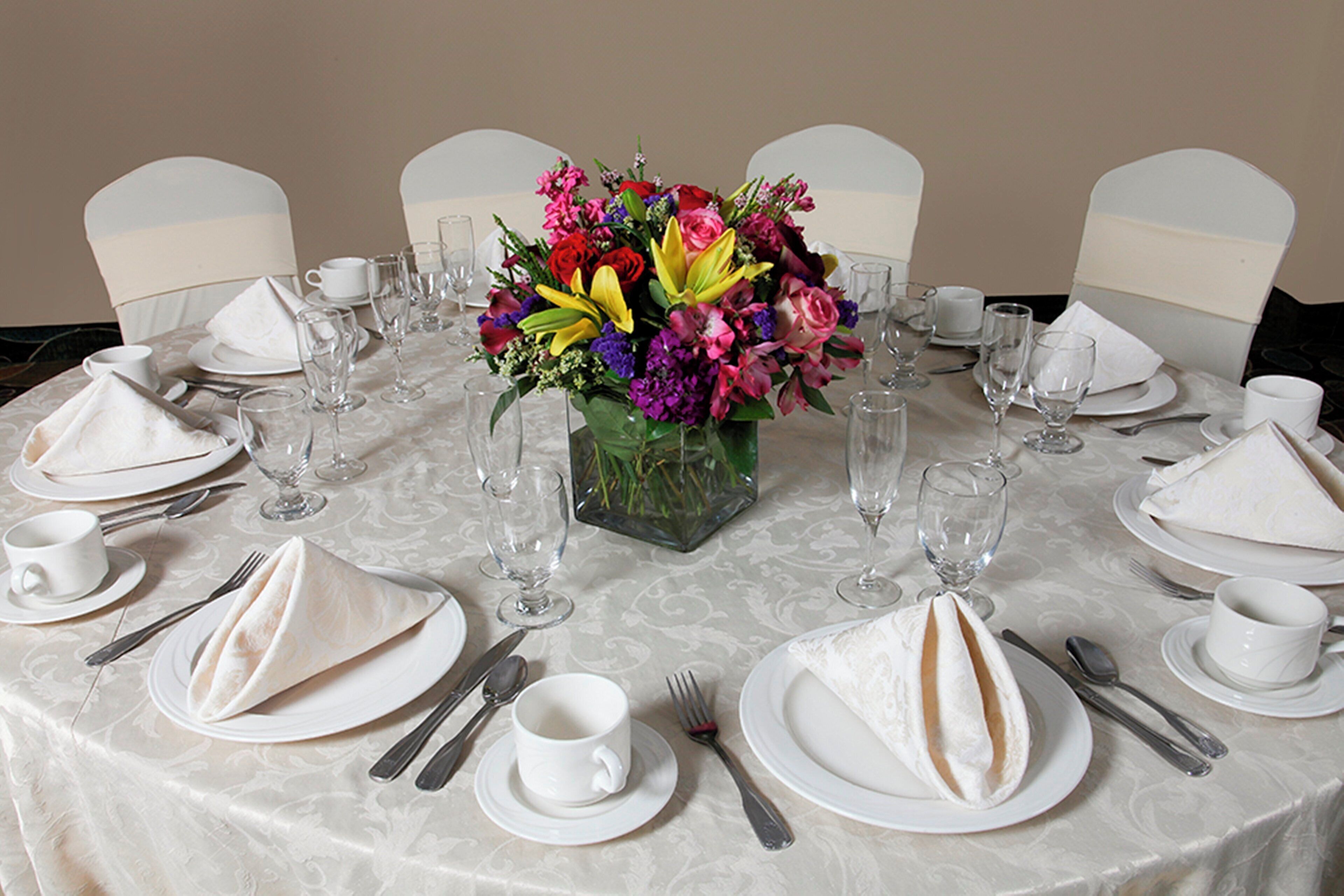 Una mesa de comedor redonda dispuesta para una comida, con platos blancos, cubiertos y vasos transparentes. En el centro, un florero con flores de colores.