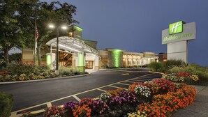 A Holiday Inn hotel with a large sign, a parking lot, and a landscaped area with colorful flowers. - Holiday Inn Westbury - Long Island by IHG (Carle Place)