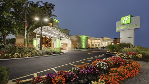A Holiday Inn hotel with a large sign, a parking lot, and a landscaped area with colorful flowers. - Holiday Inn Westbury - Long Island by IHG (Carle Place)