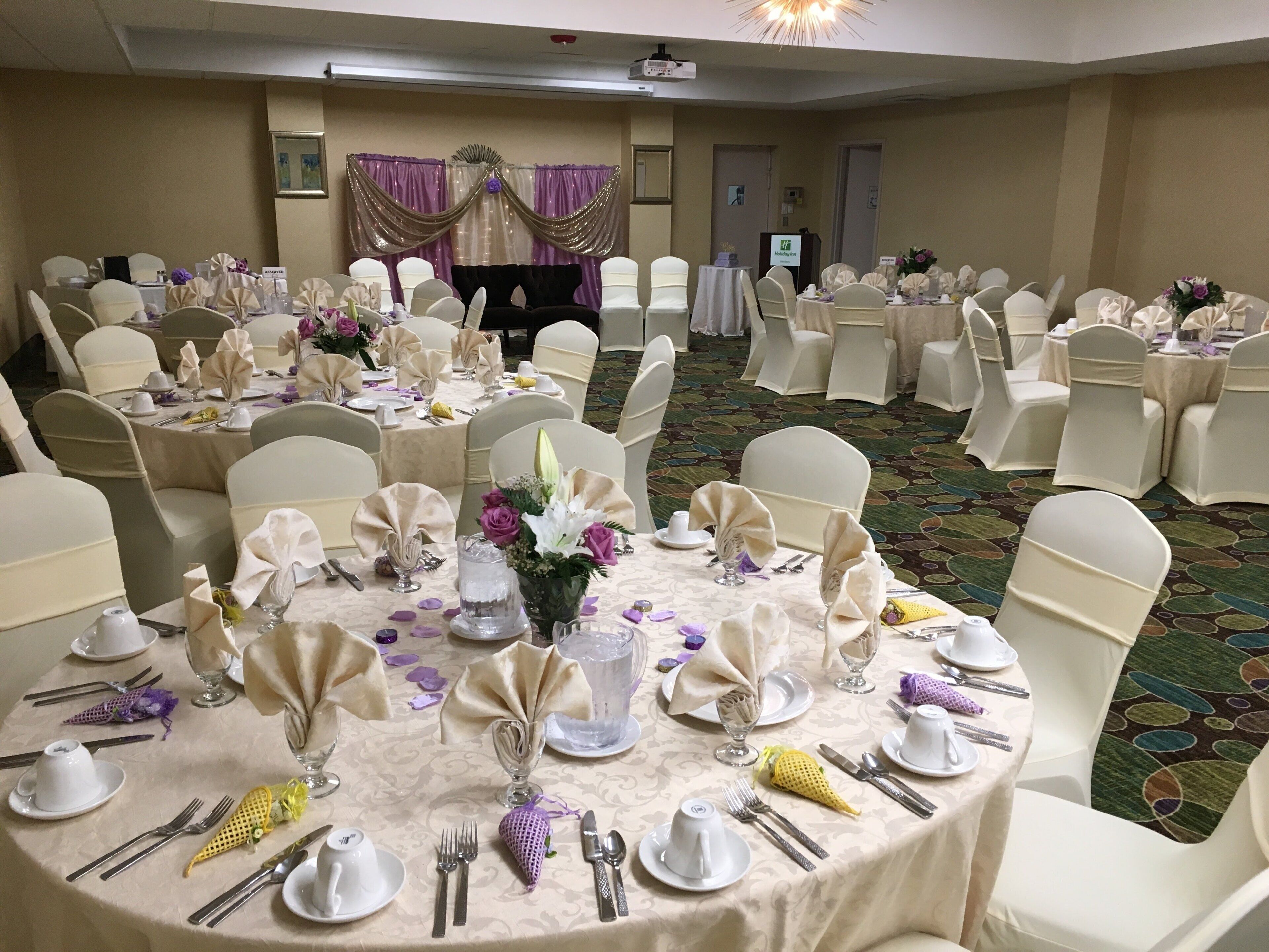 Un salón de banquetes con mesas redondas dispuestas para un evento formal, cada una con sillas blancas y vajilla.