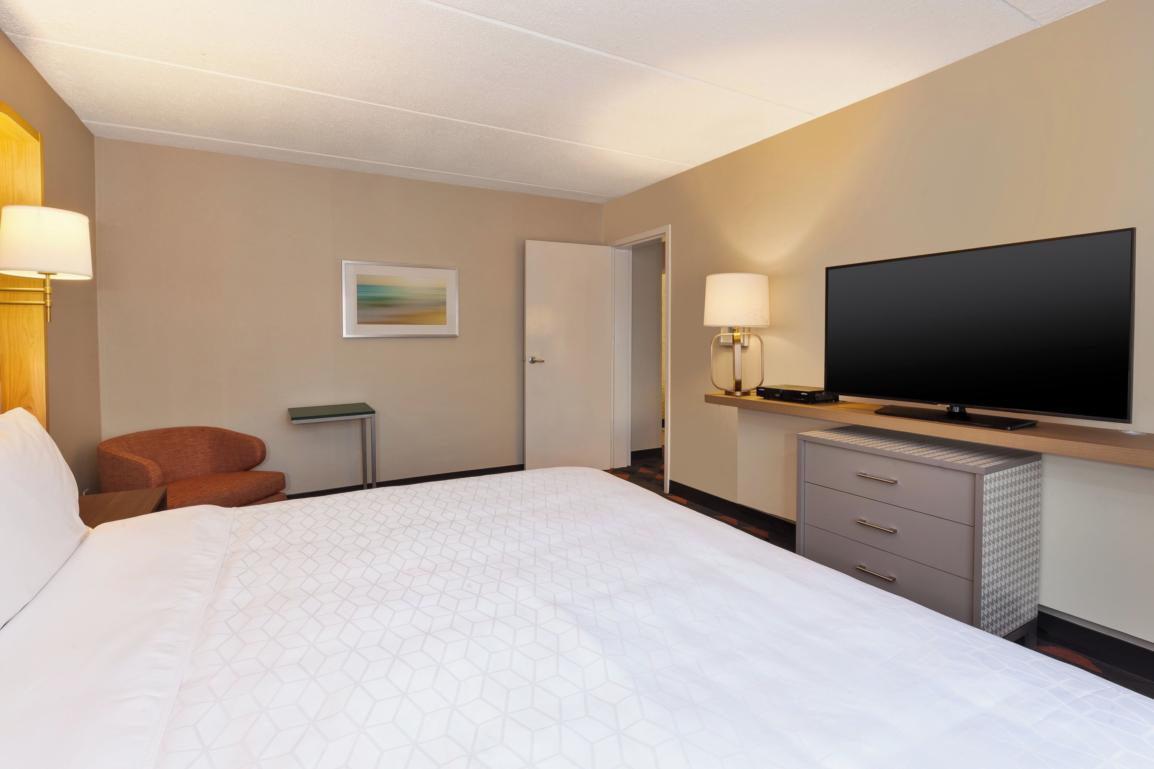Ein Hotelzimmer mit einem großen Bett, einem Fernseher, einem Schreibtisch mit Lampe und einem Sessel.