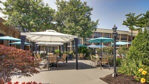 An outdoor patio area with covered seating, umbrellas, and a landscaped garden. - Holiday Inn Westbury - Long Island by IHG (Carle Place)