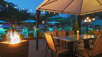 Een terras buiten met tafels en stoelen, een vuurplaats en parasols voor schaduw.